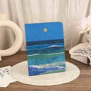 May include: A notebook with a blue cover featuring a seascape design. The artwork depicts ocean waves in shades of blue and green under a blue sky with a yellow sun. The notebook is on a woven placemat.