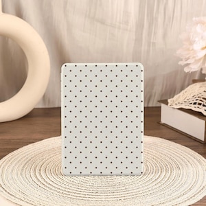 May include: A rectangular, light gray case with a repeating pattern of small, dark brown polka dots. The case is centered on a woven, circular placemat. The background includes a decorative object and a book.