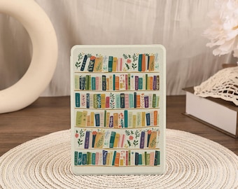 May include: A rectangular e-reader case with a light cream background featuring a repeating pattern of five horizontal shelves filled with colorful books and small green botanical illustrations interspersed between the spines of the books.