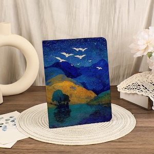 May include: A landscape-themed journal with a vibrant illustration of mountains, a lake, and flying birds. The cover features a color palette of blues, yellows, and greens, creating a serene and artistic aesthetic. The journal is displayed on a neutral-toned placemat.