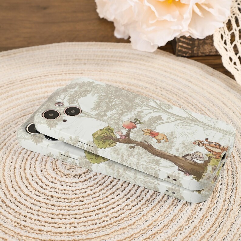 May include: Two phone cases stacked on top of each other. The cases have a light gray background with a tree design and a cartoon illustration of Winnie the Pooh and friends. The cases are on a woven mat.