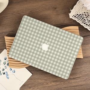 Peut inclure: Un ordinateur portable avec une housse à motif vichy gris clair et blanc. Le logo Apple est visible au centre. L'ordinateur portable repose sur un support en bois. Une carte avec le mot "NATURE" est visible en bas à gauche.