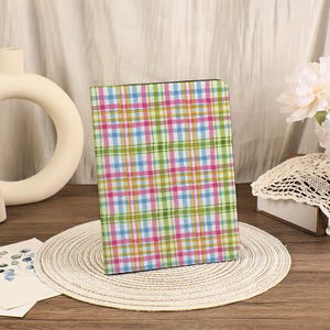 Può includere: Un quaderno a quadri colorati con un motivo di linee rosa, blu, verdi e gialle. Il quaderno è appoggiato su un sottobicchiere intrecciato. Un vaso bianco e una composizione floreale sono sullo sfondo.