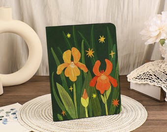 May include: A dark green notebook featuring a floral illustration of orange and yellow irises with green leaves and yellow star accents. The notebook is on a woven placemat.