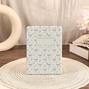 Może przedstawiać: Notatnik kompozycyjny z jasnozieloną okładką ozdobioną fioletowymi kokardkami i kwiatowymi akcentami. Na okładce znajduje się napis "COMPOSITION NOTEBOOK".