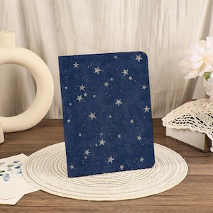 Op de afbeelding: Een marineblauw notitieboek met een sterrenhemelontwerp. De kaft heeft een patroon van witte sterren van verschillende groottes verspreid over de donkerblauwe achtergrond. Het notitieboek wordt gepresenteerd op een geweven placemat.