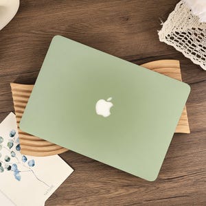 May include: A closed, sage green laptop with the Apple logo centered on the lid. The laptop rests on a wooden stand with a natural finish. A small card with a botanical illustration and the word "NATURE" is visible.