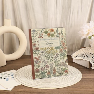 May include: A composition notebook with a cream-colored cover featuring a botanical print of various colorful flowers and plants. The center label reads Composition Notebook, Jessica, College Ruled, 120 Pages, and 8.5 x 11 inches.
