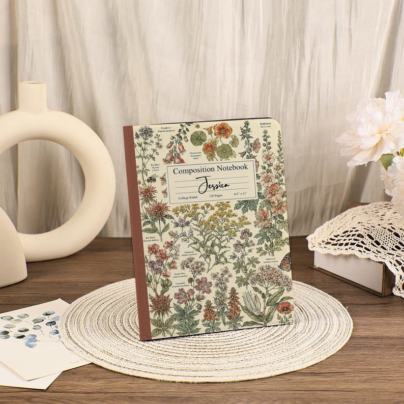 May include: A composition notebook with a cream-colored cover featuring a botanical print of various colorful flowers and plants. The center label reads Composition Notebook, Jessica, College Ruled, 120 Pages, and 8.5 x 11 inches.