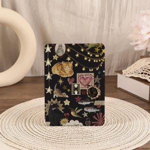 May include: A rectangular tablet case featuring a black background with a collage of illustrations including orange tabby cats, a black cat, a cat wearing a chef hat, a hummingbird, fish, hearts, stars, and string lights in gold and red tones.