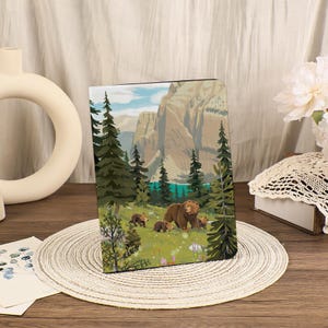 May include: A rectangular journal with a nature scene depicting a brown bear and three cubs in a forest. The background shows a lake, mountains, and evergreen trees. The journal is displayed on a woven placemat.