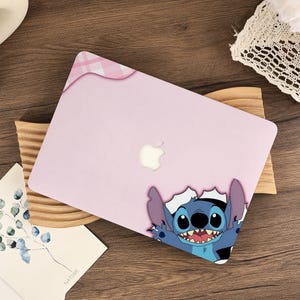 Puede incluir: Funda para portátil rosa claro con un diseño de dibujos animados de Stitch. La funda tiene un pequeño diseño de cuadros en la esquina superior izquierda y un logotipo de Apple. Stitch, de Lilo & Stitch, rompe la esquina de la funda.