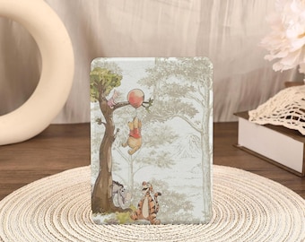May include: A rectangular e-reader cover featuring a printed illustration of Winnie the Pooh holding a red balloon, with Piglet, Eeyore, and Tigger in a forest setting. The design uses muted green and brown tones on a light background.