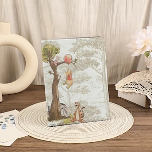 Puede incluir: Una funda para tableta de color claro con una ilustración caprichosa de Winnie the Pooh y sus amigos. La escena representa a los personajes en un entorno forestal, con Pooh sosteniendo un globo. La funda tiene un acabado mate y forma rectangular.