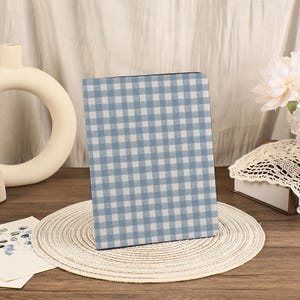 Op de afbeelding: Een notitieboek of dagboek met een blauw en wit geruit patroon. De kaft heeft een klassiek ruitpatroon in lichtblauw en wit. Het notitieboek ligt op een ronde, geweven placemat, met een witte vaas en bloemen op de achtergrond.