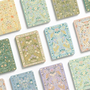 Puede incluir: Una colección de cajas metálicas rectangulares con diversos motivos florales en tonos pastel y apagados. Cada caja muestra una ilustración botánica única con pequeñas mariposas y un nombre personalizado impreso en letra cursiva al centro.