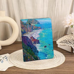 May include: A landscape painting of a coastal scene with cliffs, a beach, and ocean waves. The artwork features vibrant blues, greens, and purples, with a textured, impressionistic style. The painting is displayed on a tablet case.