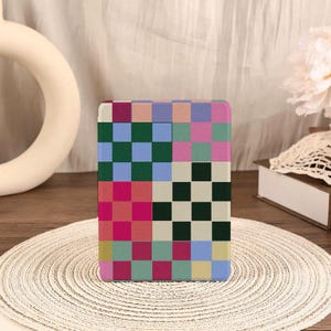 Puede incluir: Una funda rectangular para lector electrónico con un patrón de tablero de ajedrez multicolor. El diseño consta de varios bloques cuadrados en tonos verde oscuro, azul claro, magenta, rosa suave, crema y amarillo apagado en cuadrícula.