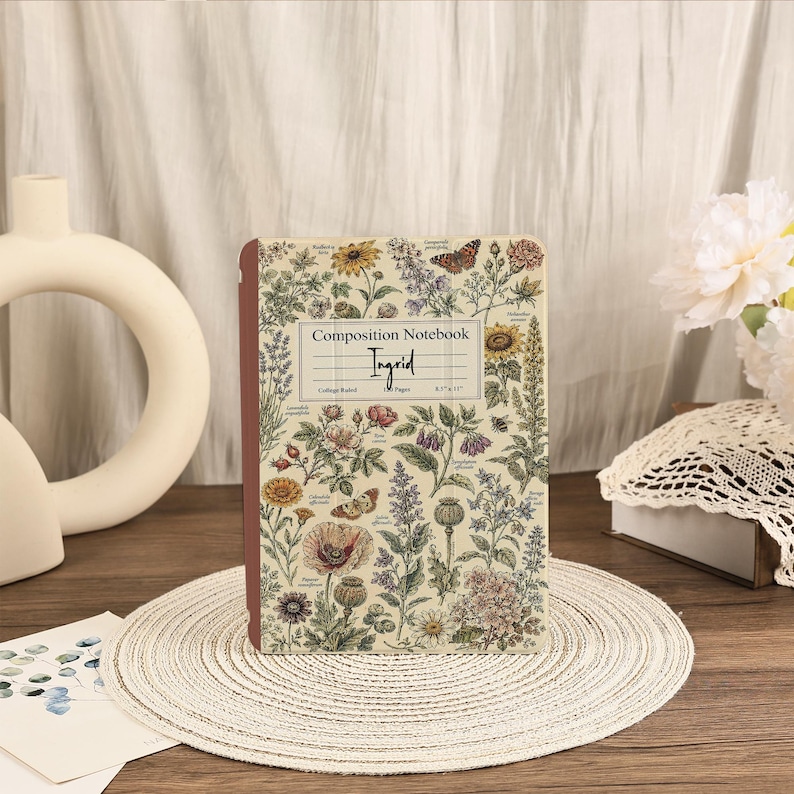 May include: A composition notebook with a cream-colored cover featuring a botanical print of various flowers, plants, and butterflies. The center label reads Composition Notebook, Ingrid, College Ruled, 100 Pages, 8.5" x 11". The spine is dark red.