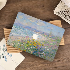 May include: A laptop skin featuring an impressionist-style painting of a coastal landscape with a sandy beach, blue ocean, and a vibrant field of yellow, pink, and purple wildflowers under a soft blue sky with clouds, centered with an Apple logo.