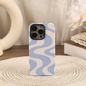 May include: A phone case in light blue and cream with a wavy pattern. The case is rectangular with rounded corners and a camera cutout. The design features flowing, curved lines in a retro style, reminiscent of the 1970s.