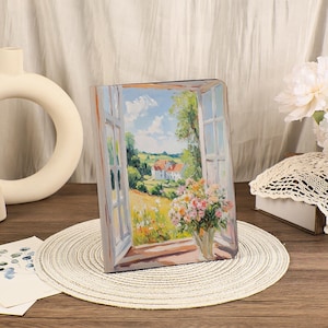 May include: A rectangular art piece showing a landscape scene through an open window. The artwork features a vase of flowers on a windowsill, with a sunny field and a house in the background. The colour palette includes blues, greens, and yellows.