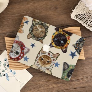 Op de afbeelding: Een laptopskin met een crèmekleurige achtergrond en een herhalend patroon van katten die verschillende mutsjes met bloemen- en fruitprints dragen. Het ontwerp bevat kleine blauwe sterren tussen de kattenillustraties en een centrale uitsparing.