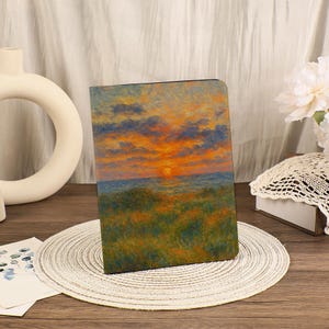 Könnte beinhalten: Ein rechteckiges Notizbuch mit einem Sonnenuntergangs-Landschaftsgemälde. Das Kunstwerk zeigt einen leuchtend orangefarbenen und gelben Himmel mit blauen Wolken über einem blauen Meer und einem grünen, grasbewachsenen Vordergrund. Das Notizbuch liegt auf einem weißen Platzset.