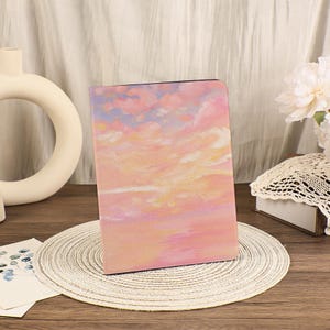 Könnte beinhalten: Ein Notizbuch mit festem Einband und Sonnenuntergangsmotiv. Der Einband zeigt ein Aquarellgemälde eines rosa, orangefarbenen und violetten Himmels mit Wolken. Das Notizbuch liegt auf einem runden, gewebten Platzset.