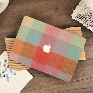 Puede incluir: Una funda de portátil colorida con un diseño de cuadros en tonos naranja, rosa, azul y verde. La funda presenta el logotipo de Apple en blanco. Descansa sobre un soporte de madera, con una tarjeta que dice "NATURE" en la esquina.