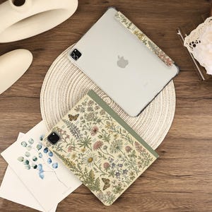 May include: A tablet case featuring a botanical illustration of wildflowers, herbs, and butterflies in muted earth tones on a cream background, paired with a clear tablet case that has a matching floral strip along the side edge.