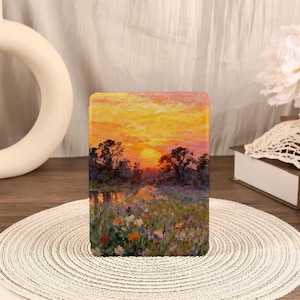 May include: A rectangular case featuring a landscape painting of a sunset over a body of water. The artwork depicts a vibrant sky with orange and yellow hues, silhouetted trees, and colorful wildflowers in the foreground. The case is resting on a woven placemat.