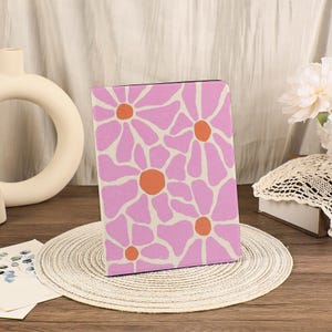 Könnte beinhalten: Ein rechteckiges Notizbuch oder Journal mit einem rosa-orangenen Blumenmuster. Das Design zeigt große, stilisierte Blumen mit rosa Blütenblättern und orangefarbenen Zentren auf cremefarbenem Hintergrund. Das Notizbuch wird auf einem gewebten Platzset präsentiert.