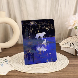 May include: A notebook with a fantasy illustration of two white wolves on a blue and gold landscape under a starry night sky. The cover features a reflective water effect, with the wolves' reflections visible.