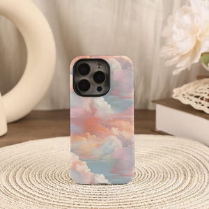 Puede incluir: Funda de teléfono inteligente con un diseño de nubes pastel en tonos rosa, naranja y azul. La funda tiene un acabado brillante y un recorte para la cámara. La estética general es suave y soñadora.