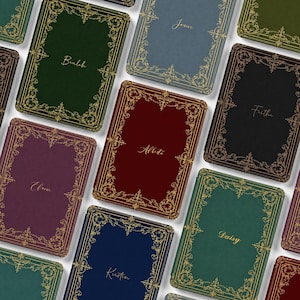 May include: A collection of rectangular journals in various colours including dark green, blue, burgundy, purple, and black. Each journal features an ornate gold filigree border and a unique name printed in gold script in the centre of the cover.