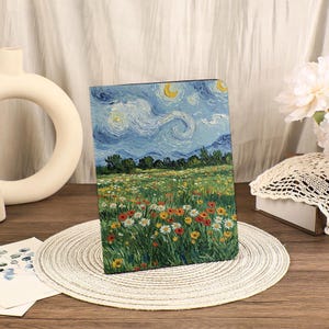 Puede incluir: Un cuaderno rectangular con una pintura de un cielo estrellado, una luna creciente y un campo de flores coloridas, al estilo de Van Gogh. La obra de arte presenta nubes arremolinadas y un prado vibrante. El cuaderno se muestra sobre un salvamanteles tejido.