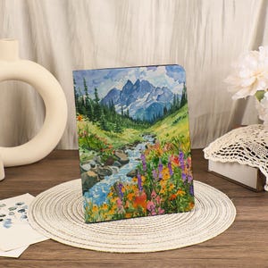 May include: A watercolour painting depicting a mountain scene with a stream and vibrant wildflowers. The artwork showcases a blue sky, snow-covered mountains, and verdant green trees. The painting is presented on a decorative item.