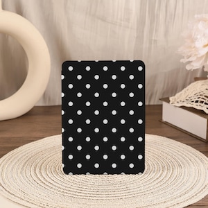 May include: A black rectangular object with a white polka dot pattern. The object is standing upright on a round, woven placemat. The background includes a white floral arrangement and a decorative cream-colored object.