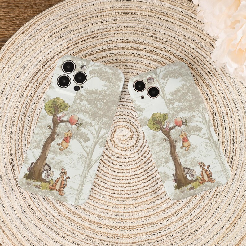 May include: Two phone cases with a whimsical forest scene featuring cartoon characters. The cases have a light beige background with a repeating tree pattern. The cartoon characters are in the foreground, with a tree and a red balloon. The cases are on a beige woven surface.