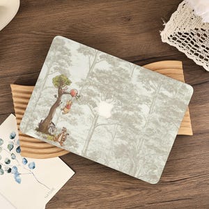 Peut inclure: Un autocollant pour ordinateur portable illustrant une forêt vert clair avec les personnages de Winnie l'ourson, dont Winnie, Tigrou et Bourriquet. Le motif montre Winnie tenant un ballon rouge près d'un arbre, avec une découpe centrale.