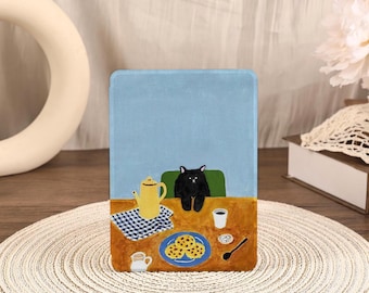 Puede incluir: Una funda azul para tableta con una ilustración de un gato negro sentado en una mesa. La mesa tiene una tetera, galletas y café. El fondo es azul claro. Diseño caprichoso.