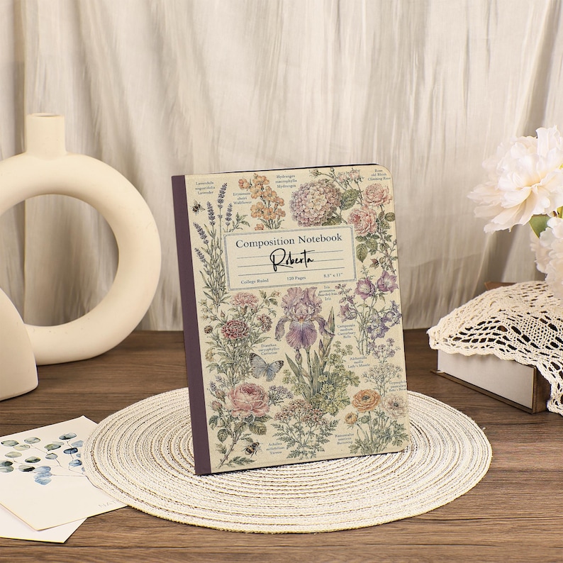 May include: A composition notebook with a cream-colored cover featuring a botanical illustration of various flowers, plants, and a butterfly. The text reads Composition Notebook, Roberta, College Ruled, 128 Pages, and 8.5 x 11 inches.