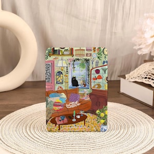 Puede incluir: Una impresión artística rectangular que representa una acogedora escena de sala de estar. La ilustración presenta un sofá marrón, un gato negro y varios elementos decorativos, incluyendo una mesa de centro y un armario. La paleta de colores incluye tonos cálidos y detalles vibrantes.