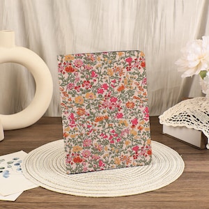 Puede incluir: Funda rectangular para tableta con estampado floral. El diseño presenta flores rosas, naranjas y amarillas sobre un fondo beige claro. La funda es de tela y tiene un estilo de estampado botánico vintage. Diseñada para proteger una tableta.