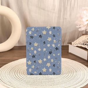 May include: A rectangular, blue case with a pattern of gold, silver, and dark blue stars. The stars are of varying sizes and are scattered across the surface. The case is sitting on a white, woven placemat.