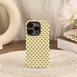 May include: A pale yellow smartphone case featuring a repeating pattern of small, evenly spaced dark brown polka dots. The case includes a black rectangular camera cutout at the top left corner, designed to accommodate a multi-lens camera system.