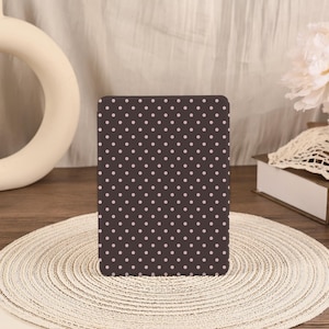 May include: A rectangular electronic reader device featuring a dark brown exterior surface covered in a repeating pattern of small, light pink polka dots. The device is positioned vertically, displaying a uniform grid of dots across its entire front face.