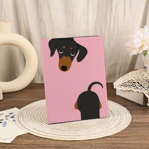 May include: Pink tablet case featuring a black and brown dachshund dog illustration. The dog's head and tail are visible on the case. The case is on a woven placemat on a wooden surface.