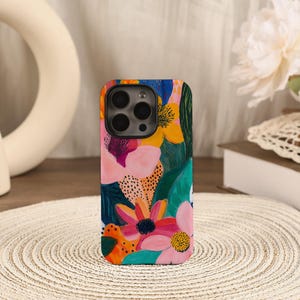 Peut inclure: Une coque de smartphone rectangulaire ornée d'un motif floral aux couleurs vives. Le design présente des fleurs peintes dans des tons de rose, orange et jaune, rehaussées par un feuillage vert profond et des petits points noirs sur fond bleu.